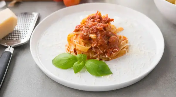Talerz z makaronem tagliatelle polanym sosem pomidorowym, posypany parmezanem i udekorowany bazylią.
