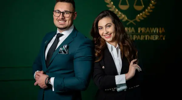 Para prawników w eleganckich garniturach na tle zielonej ściany z logo kancelarii.