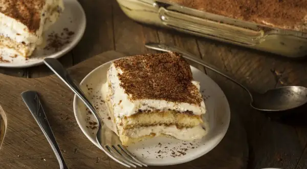 Une part de tiramisu, dessert dont l'origine est débattue, sur une assiette avec une fourchette. Le dessert est saupoudré de cacao.
