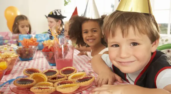 Glada barn med kalashattar vid ett dukat bord med tårtor och frukter. Perfekt för kalaslekar 5 år!