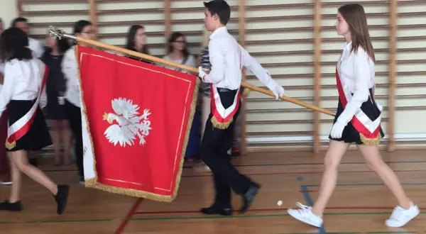 Uczniowie w galowych strojach tworzą poczet sztandarowy, niosąc biało-czerwony sztandar z godłem Polski.