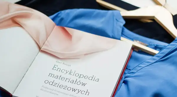 Książka "Encyklopedia materiałów odzieżowych" leży na niebieskiej i czarnej tkaninie. Może zawierać informacje o wadach wiskozy.