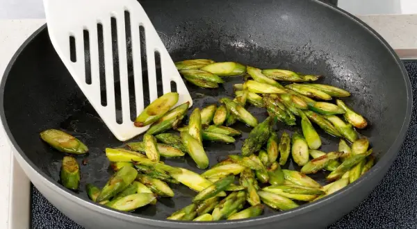 Geschnittener Spargel brät in einer Pfanne. Ein weißer Pfannenwender bewegt die grünen Stücke.