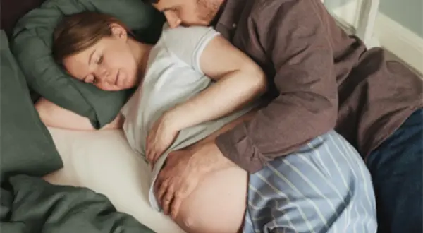 En man kramar om sin gravida partner. Hon vilar huvudet på en kudde. Kanske en bild från 1177 om hinnsvepning.