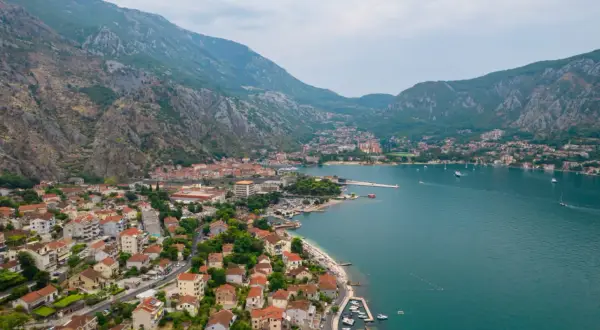 Widok na Zatokę Kotorską z górami w tle. Widać miasto z czerwonymi dachami, plażę i łodzie. Kotor atrakcje zachwycają!
