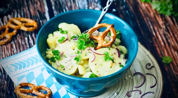Klassisches Rezept Kartoffelsalat mit roten Zwiebeln und Petersilie, garniert mit einem Brezel.