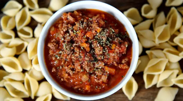 Herzhaftes Bolognese-Rezept mit Hackfleisch und Tomaten, garniert mit Kräutern, umgeben von ungekochten Nudeln.