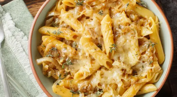 Makaron penne z karmelizowaną cebulą i roztopionym serem, udekorowany ziołami.