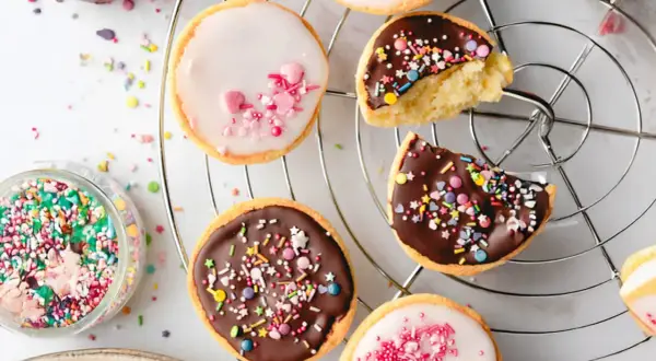 Bunte mini Amerikaner mit Schokoladen- und Zuckerguss, bestreut mit Streuseln und Herzen, auf einem Kuchengitter.
