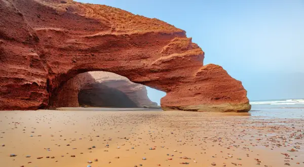 Skalny łuk na piaszczystej plaży w Maroku. Słońce wdziera się do jaskini, tworząc magiczną atmosferę.