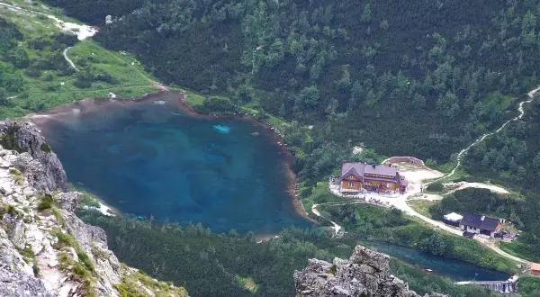 Widok na zielony staw kieżmarski otoczony lasem i górami. W pobliżu schronisko i ścieżki.