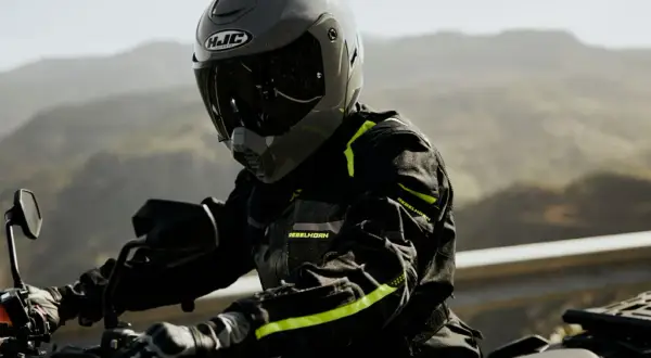 Motocyklista w szarym kasku HJC i czarnej kurtce z neonowymi detalami jedzie motocyklem po górskiej drodze.