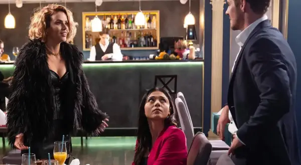 Izak Ikusia w czarnym futrze i skórzanym gorsecie, rozmawia z parą przy stoliku w restauracji.