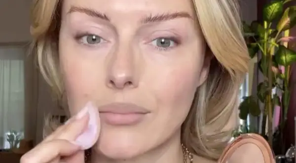 Eine Frau trägt mit einem Schwämmchen puder auf ihr Gesicht auf. Ihr blondes Haar umrahmt ihr Gesicht, während sie sich auf ihr Make-up konzentriert.