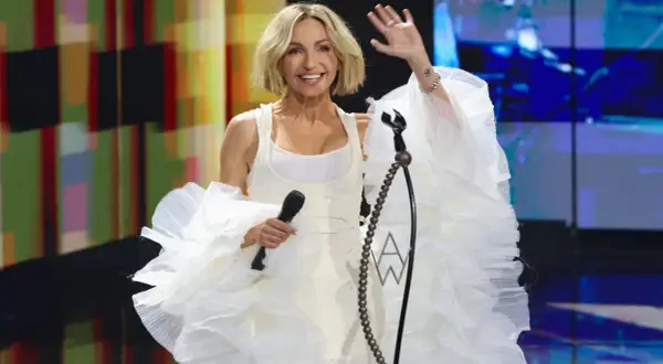 Anna Wyszkoni w białej, falbaniastej kreacji pozdrawia publiczność. W ręku trzyma mikrofon, gotowa do wykonania swoich utworów.