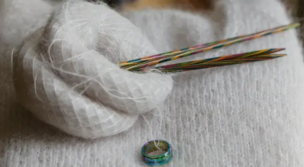 Ein Knäuel weiches, flauschiges Mohairgarn liegt neben bunten Stricknadeln auf einem Strickstück. Was ist Mohair? Es ist eine luxuriöse Faser.