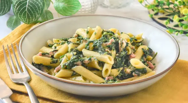 Un plato de pasta penne con espinacas y trocitos de jamón, servido en un cuenco sobre una servilleta amarilla.