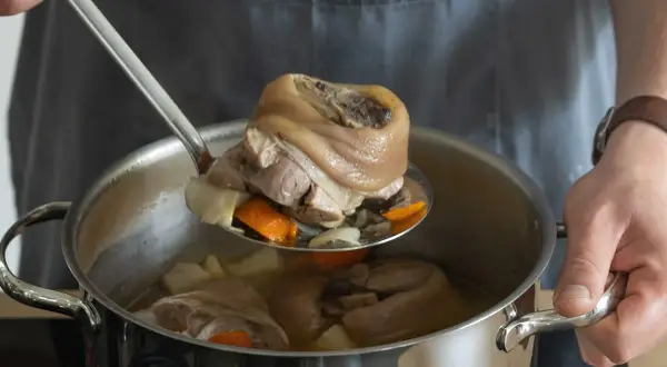 Eisbein zubereiten im Topf: Ein Koch hebt mit einer Schöpfkelle ein gekochtes Eisbein aus einem Topf mit Gemüse und Brühe.