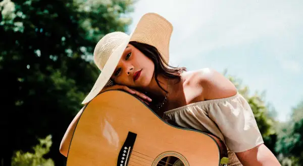 Kobieta w kapeluszu opiera się o gitarę, marząc o najpiękniejszych piosenkach na gitarę.