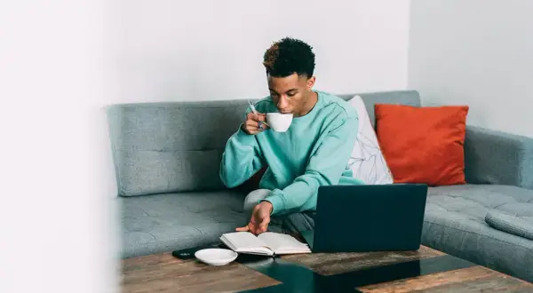 Ung man sitter i soffan med en bok och en kopp kaffe. Han tar en klunk tein koffein medan han läser.