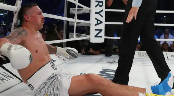 Boxer Oleksandr Usyk sits on the canvas, leaning against the ropes, appearing to recover from a usyk low blow. The referee stands nearby.