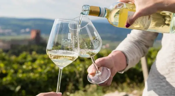 Versando vino bianco in calici con logo Soave, sullo sfondo un paesaggio collinare con vigneti e un castello. Un brindisi al Soave Superiore DOCG.
