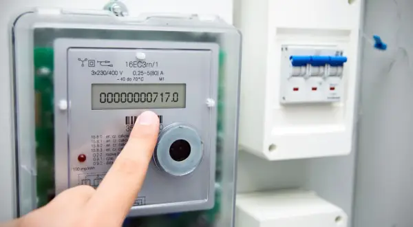 Palec wskazuje licznik energii elektrycznej, pokazujący 717.0 kilowatogodzina. Obok widoczny jest wyłącznik nadprądowy.