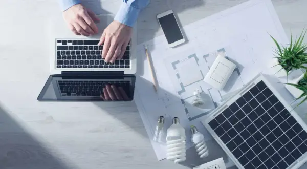Analiza sprawności paneli fotowoltaicznych na laptopie. Na biurku projekt, żarówki, gniazdko i telefon.