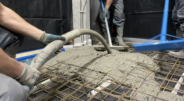 Pracownicy wykonują pompowanie betonu na zbrojenie. Beton płynie z węża, a drugi pracownik rozprowadza go łopatą.