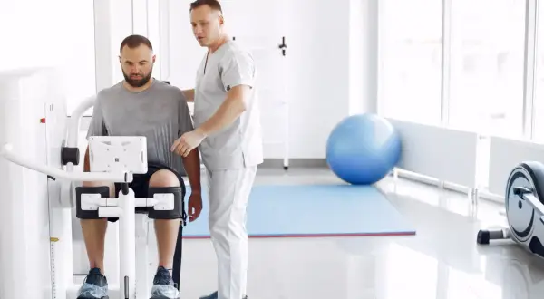 Fizjoterapeuta pomaga pacjentowi na maszynie rehabilitacyjnej w jasnym gabinecie.