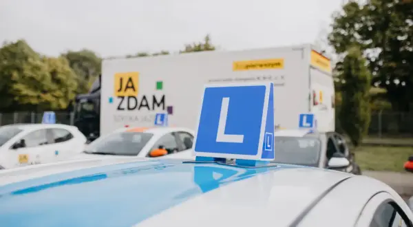 Na dachu białego samochodu nauki jazdy stoi niebieska tablica z literą "L". W tle ciężarówka z napisem "JA ZDAM". Ile można mieć błędów na egzaminie teoretycznym?