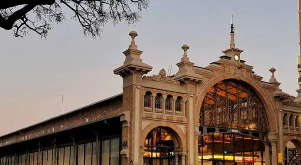El **mercado central Zaragoza** con su arquitectura de hierro y cristal, iluminado al atardecer. Gente pasea por la entrada principal.