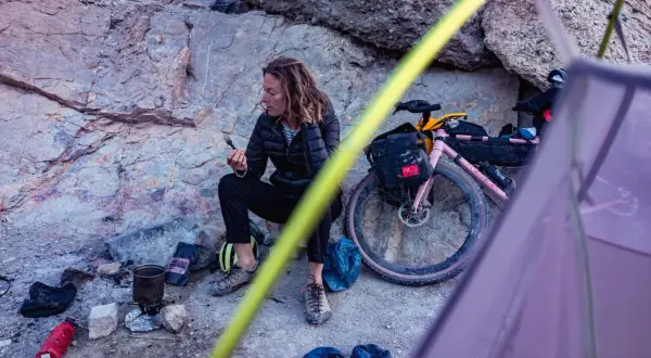 Pause sur une sortie gravel : une cycliste se repose près de son vélo rose, une tasse à la main, au milieu d'un paysage rocheux.