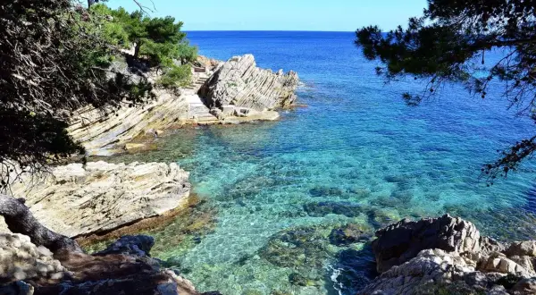 Agua cristalina en una cala rocosa, con escaleras de piedra que bajan al mar. El sol ilumina el fondo marino, creando un espectáculo de luz y color.