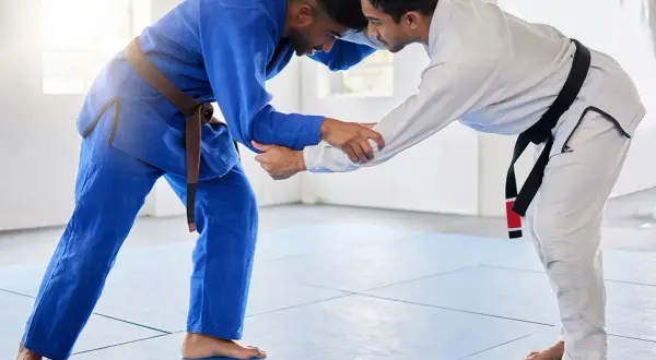 Dwóch mężczyzn w kimonach do BJJ, jeden w niebieskim z brązowym pasem, drugi w białym z czarnym pasem, ćwiczy chwyty na macie.