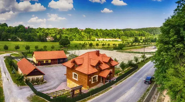 Drewniane budynki rancza z czerwonymi dachami, otoczone zielenią, pod błękitnym niebem z chmurami.