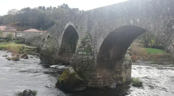 El histórico puente Maceira cruza un río caudaloso, con casas de tejados rojos y árboles frondosos al fondo.