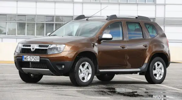 Brązowy Dacia Duster na parkingu, gotowy do testu. Widoczny kierowca za kierownicą.