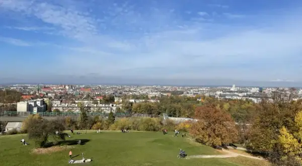 Widok na Kraków z zielonego wzgórza, gdzie ludzie odpoczywają. W oddali rozciąga się panorama miasta, a jesienne drzewa dodają uroku. To idealne miejsce na spacer, jak na **Kopcu Kraka**.