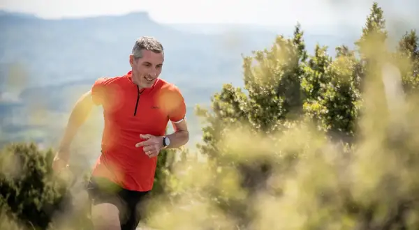 Un homme en tenue de trail run, souriant, court sur un sentier de montagne.