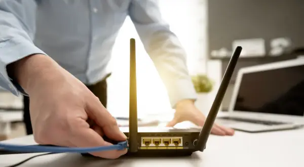 Dłoń podłącza kabel do routera, co może być pierwszym krokiem, jak zresetować router. W tle widać laptopa.