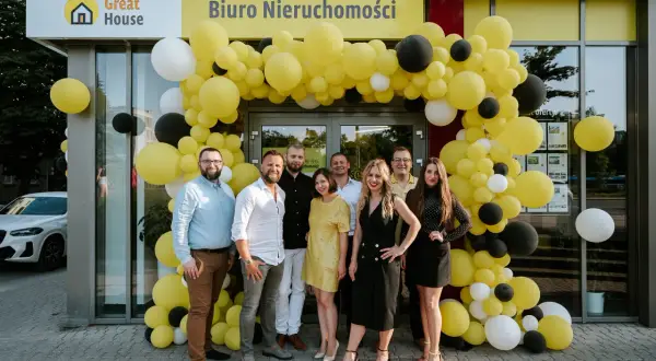 Grupa ludzi pozuje przed biurem nieruchomości ozdobionym żółtymi balonami.