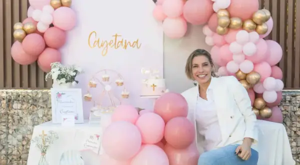 Una mujer sonríe junto a una mesa decorada para una comunión casera, con globos rosas y dorados, un pastel y un cartel de bienvenida.