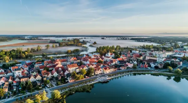Poranna mgła nad rzeką i polami, otaczająca urokliwe miasteczko z czerwonymi dachami. To prawdziwa kraina 100 jezior, gdzie spokój miesza się z pięknem natury.