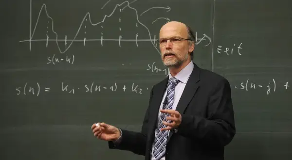 Profesor w garniturze tłumaczy równania na tablicy. Zastanawiamy się, ile godzin ma nauczyciel na etacie, gdy widzi się jego zaangażowanie.