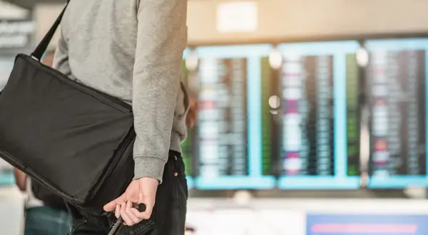 Mann mit schwarzer Reisetasche und Koffer vor Anzeigetafel am Flughafen.