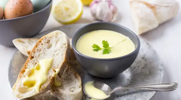 Kremowy sos aioli w miseczce, z pieczywem, jajkami, cytryną i czosnkiem w tle.