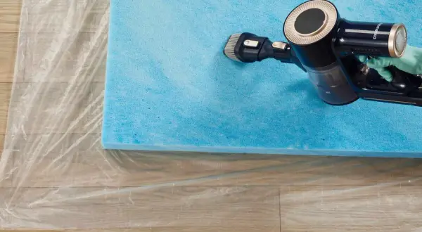 A gloved hand uses a vacuum cleaner with a brush attachment to clean a blue foam mattress topper laid on plastic sheeting.