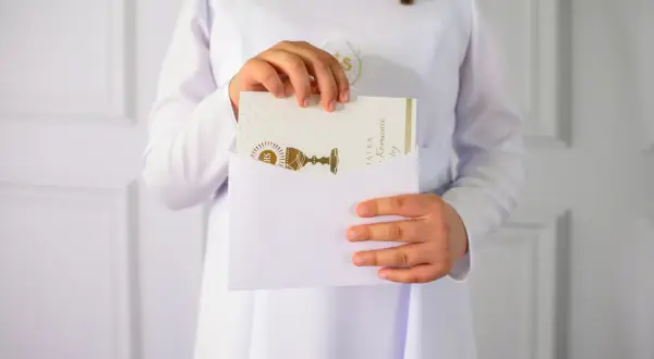 Dziecko w bieli wkłada kartkę z kielichem do koperty. Idealne na krótkie życzenia komunijne.