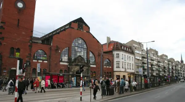 Menschen warten an einer Straßenbahnhaltestelle vor der imposanten Markthalle Breslau.